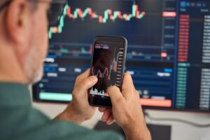 Investor reviewing stock market charts on a smartphone in front of a trading screen